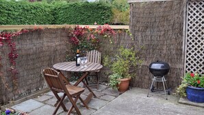 A patio with a round wooden table and two chairs, a bottle of wine, and a grill. - 1 bedroom accommodation in Rye (Rye)