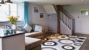 A modern living room with a sofa, a coffee table, and a vase with flowers. There is a staircase with a wooden railing leading to another level. - 2 bedroom accommodation in St Neot, near Liskeard (St Neot, near Liskeard)