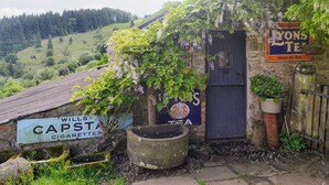 Un bâtiment rustique avec une porte sombre, des enseignes pour les cigarettes « Wills » et le thé « Yons », et une vue sur une vallée verdoyante.