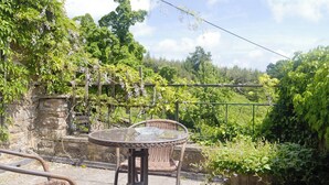 Une terrasse extérieure avec une table ronde en verre et des chaises en osier, entourée d’une végétation luxuriante et d’un mur de pierre.