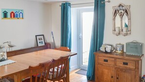 A dining area with a wooden table and chairs, a cabinet with a 'BREAD' sign, a framed picture on the wall, and a window with curtains.