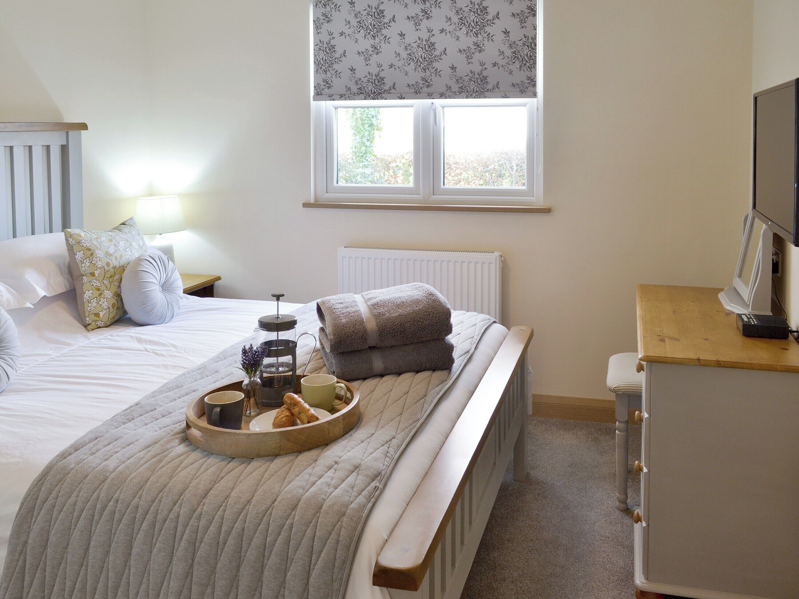 A neatly made bed with a tray of breakfast items, a bedside table with a lamp, and a window with a floral blind.