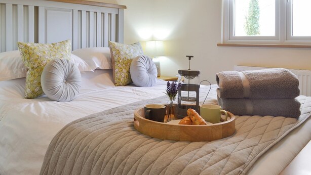 Ein ordentlich bezogenes Bett mit floralen Kissen, ein Nachttisch mit Lampe, eine Ablage mit Croissants und ein Fenster mit Blick ins Grüne.