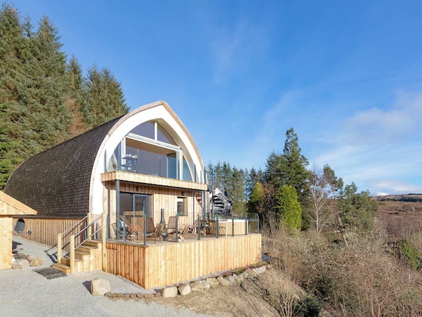 A wooden cabin with a large glass window, wooden deck, and a stone pathway leading to the entrance.