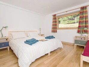 A neatly made bed with striped bedding, a bedside table with a lamp, and a window with curtains. - Jill Strawbale House- UKC2935 (Strontian, near Fort William)