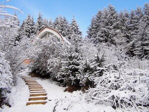 A snow-covered pathway leading to a building with a triangular roof, surrounded by dense evergreen trees. - Jill Strawbale House- UKC2935 (Strontian, near Fort William)