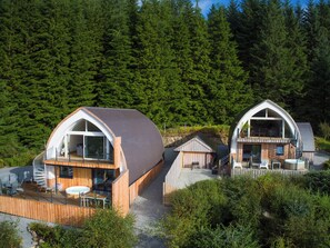 Twee moderne vakantiehuisjes met gebogen daken, houten terrassen en glazen deuren, omgeven door dicht bos.