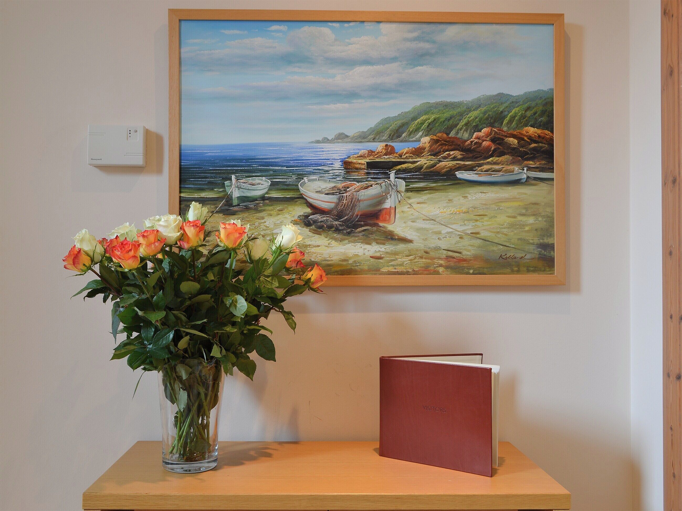 A painting of boats on a beach, a vase of flowers, and a red book on a wooden table.