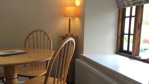 A dining area with a round wooden table and four wooden chairs. A table lamp is on the wall beside the table. A window with blinds is on the wall to the right. - Millstones is a lovely property for two. (Seatown, Chideock)