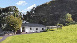 Une petite maison blanche avec un mur de pierres, une clôture en bois et une colline couverte d’herbe.