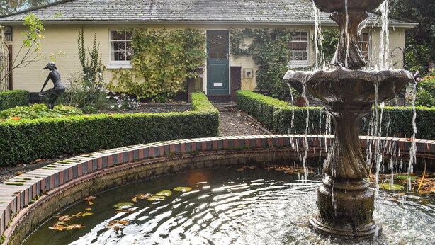 A garden with a fountain, a hedge, and a building with a green door.