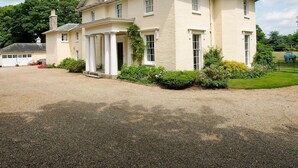 A large, cream-colored building with a symmetrical facade, a central entrance with columns, and a gravel driveway.