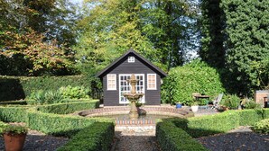 A small, dark-colored house with a central fountain, surrounded by well-manicured hedges and a gravel path.