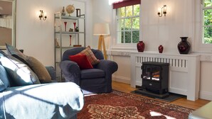 A cozy living room with a fireplace, a blue armchair, a patterned rug, and a bookshelf with decorative items.