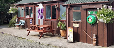 Ein Holzgebäude mit einem grünen Dach, einem Picknicktisch und einer Bank. Schilder weisen auf Toiletten und einen Defibrillator hin.