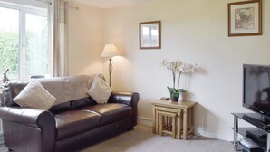 A living room with a leather sofa, a standing lamp, a small table with a plant, a framed picture, and a television. - 3 bedroom accommodation in Frostenden, near Beccles (Frostenden, near Beccles)
