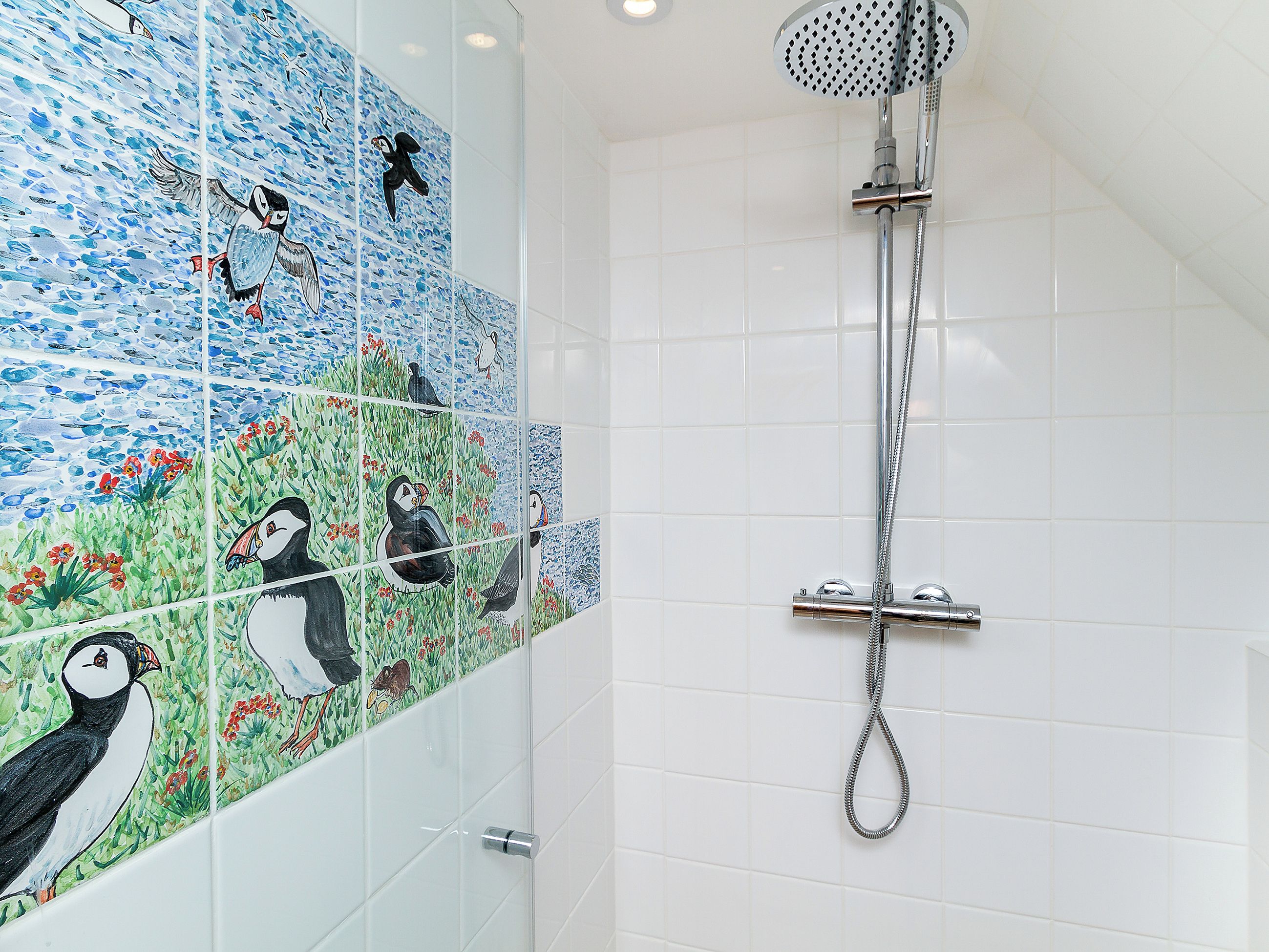 A bathroom with a shower, white tiled walls, and a mosaic wall with bird illustrations.