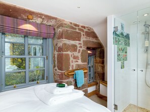 A bedroom with a large bed, a stone wall, a window with blinds, and a shower area with a glass door.