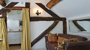 A rustic interior with exposed wooden beams, a brown leather sofa with patterned cushions, a kitchen area, and a window with a mirror. - 1 bedroom accommodation in Betley, near Nantwich (Betley, near Nantwich)