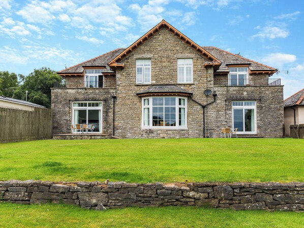 A two-story stone house with a stone wall in front, large windows, and a sloped roof.