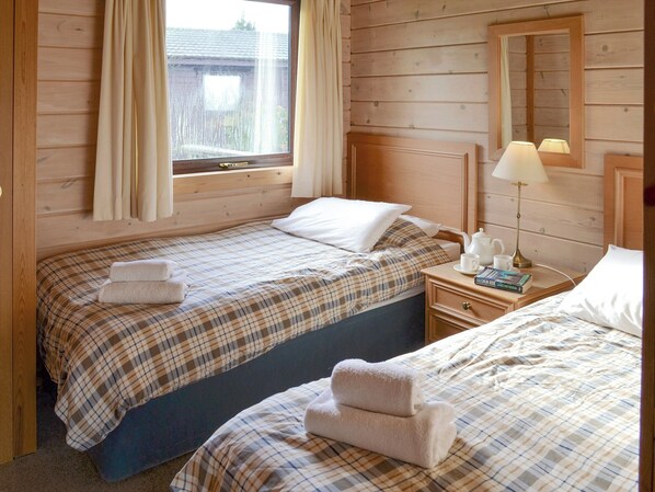 Una habitación con dos camas, cada una con cubrecama a cuadros y almohadas blancas. Hay una mesita de noche con una lámpara, un libro y una taza. La habitación tiene paredes de madera y una ventana con cortinas.
