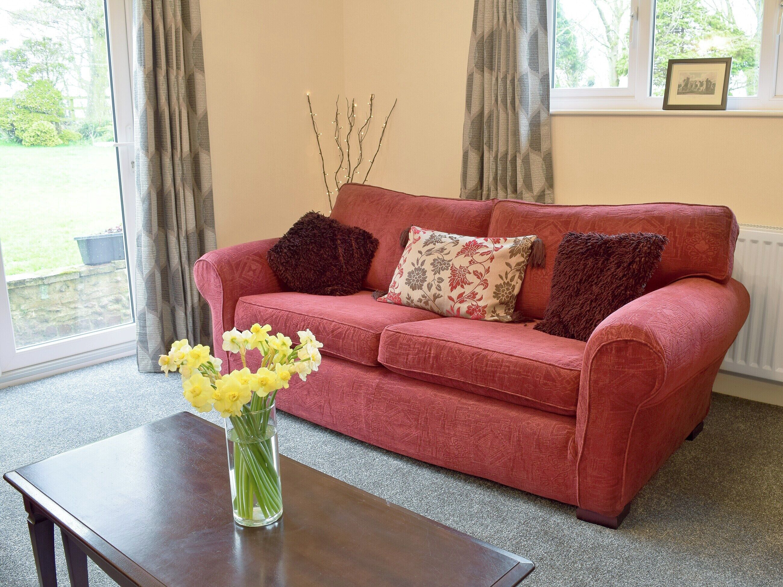 A red sofa with floral cushions, a vase with yellow flowers, and a window with curtains.