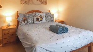 A neatly made bed with a white bedspread and a blue towel, two bedside lamps, and a painting of a beach scene on the wall. - 1 bedroom accommodation in Mount Hawke (Mount Hawke)