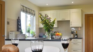 Una cocina con armarios blancos, una ventana con persianas, un jarrón con flores, un cuenco de frutas y dos copas de vino tinto sobre la encimera.