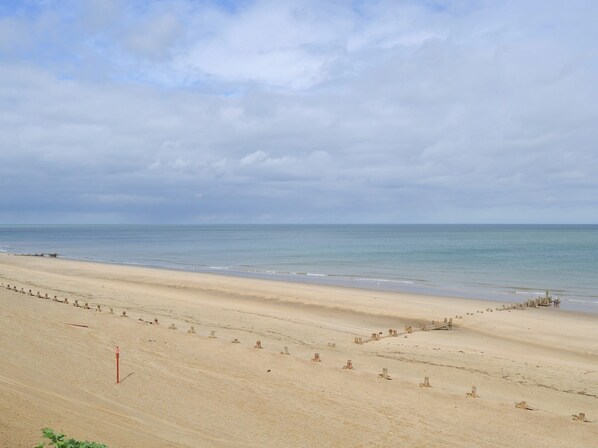 A wide sandy beach with a calm sea, a cloudy sky, and a few scattered structures along the shore.