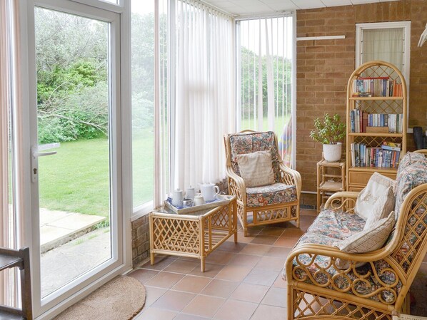 A sunlit room with wicker furniture, a bookshelf, and a view of a green garden. - 3 bedroom accommodation in Bacton, near Happisburgh (Bacton, near Happisburgh)