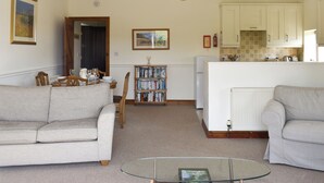 A living room with a sofa, a glass coffee table, a bookshelf, a kitchen area with cabinets, and a chandelier. - 2 bedroom accommodation in Thorncombe, near Broadwindsor (Thorncombe, near Broadwindsor)