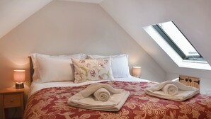 Un dormitorio con techo inclinado, un tragaluz, una cama con ropa de cama floral, dos lámparas de noche y toallas ordenadas sobre la cama.