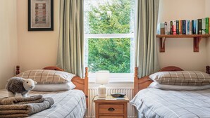 Een slaapkamer met twee bedden, een nachtkastje, een lamp, een boekenkast en een raam met uitzicht op bomen.