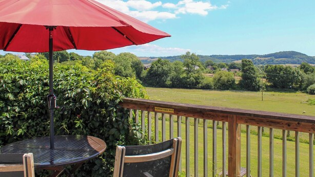 Une terrasse avec un parasol rouge, une table et des chaises, donnant sur un champ verdoyant et des collines.