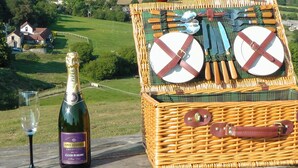 Un panier de pique-nique en osier contenant des couverts et des assiettes, une bouteille de vin et un verre, le tout posé sur une étagère surplombant un paysage rural.