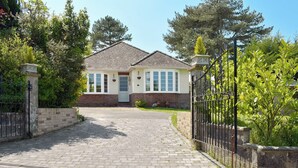 A house with a paved driveway leading to a gated entrance, surrounded by greenery and trees.