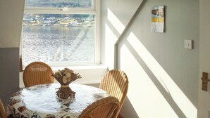Une table à manger recouverte d’une nappe aux motifs de café, des chaises en osier et un centre de table décoratif. Une fenêtre offre une vue sur un plan d’eau et des bâtiments.