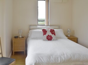 A neatly made bed with white bedding and red floral pillows, two bedside lamps, a wooden nightstand, a chair, and a window with a view of the outdoors.