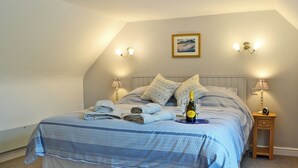 Un lit bien fait, recouvert d’une couvre-lit à motifs bleus et blancs, avec deux serviettes enroulées, une bouteille de champagne et deux verres posés sur un plateau. Deux lampes murales et un tableau encadré sont fixés au mur au-dessus du lit.