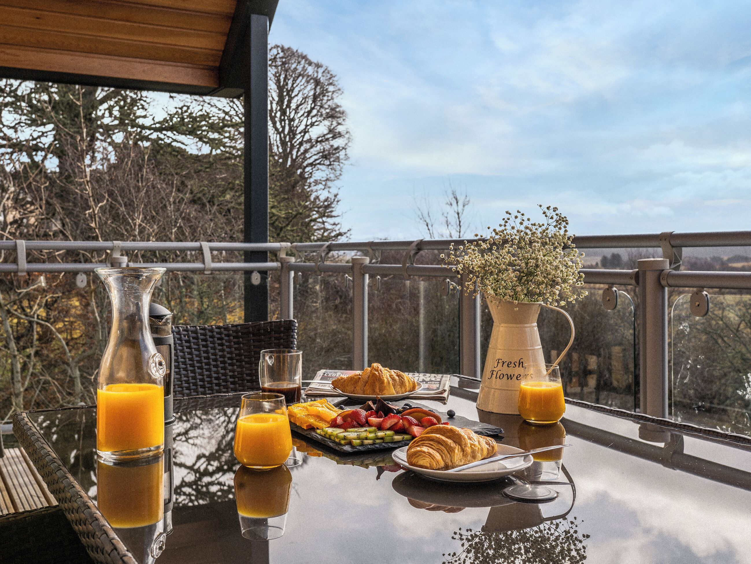 Une table de petit-déjeuner avec un croissant, des fruits, du jus et un vase de fleurs, le tout placé sur un balcon donnant sur des arbres.