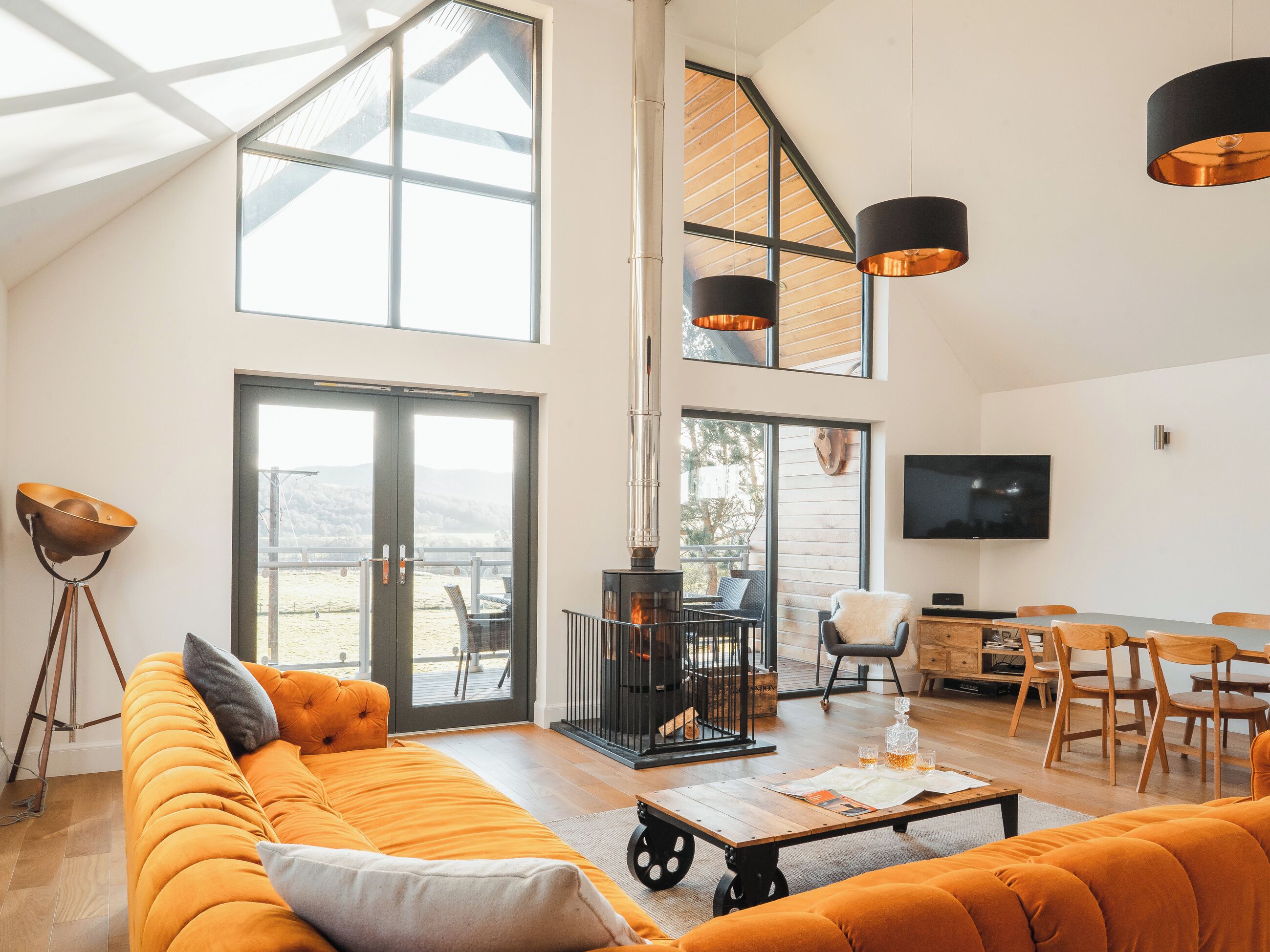 Uma sala de estar ampla com um sofá grande, uma mesa de centro e uma lareira. Há uma televisão na parede e uma varanda com vista para o exterior.