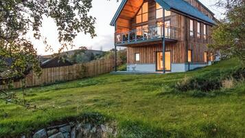 A modern wooden house with a balcony, surrounded by a grassy yard and a stone wall.