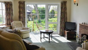 A living room with a sofa, armchair, TV, fireplace, and a view through large glass doors to a garden. - 1 bedroom accommodation in Dalwood, near Axminster (Dalwood, near Axminster)