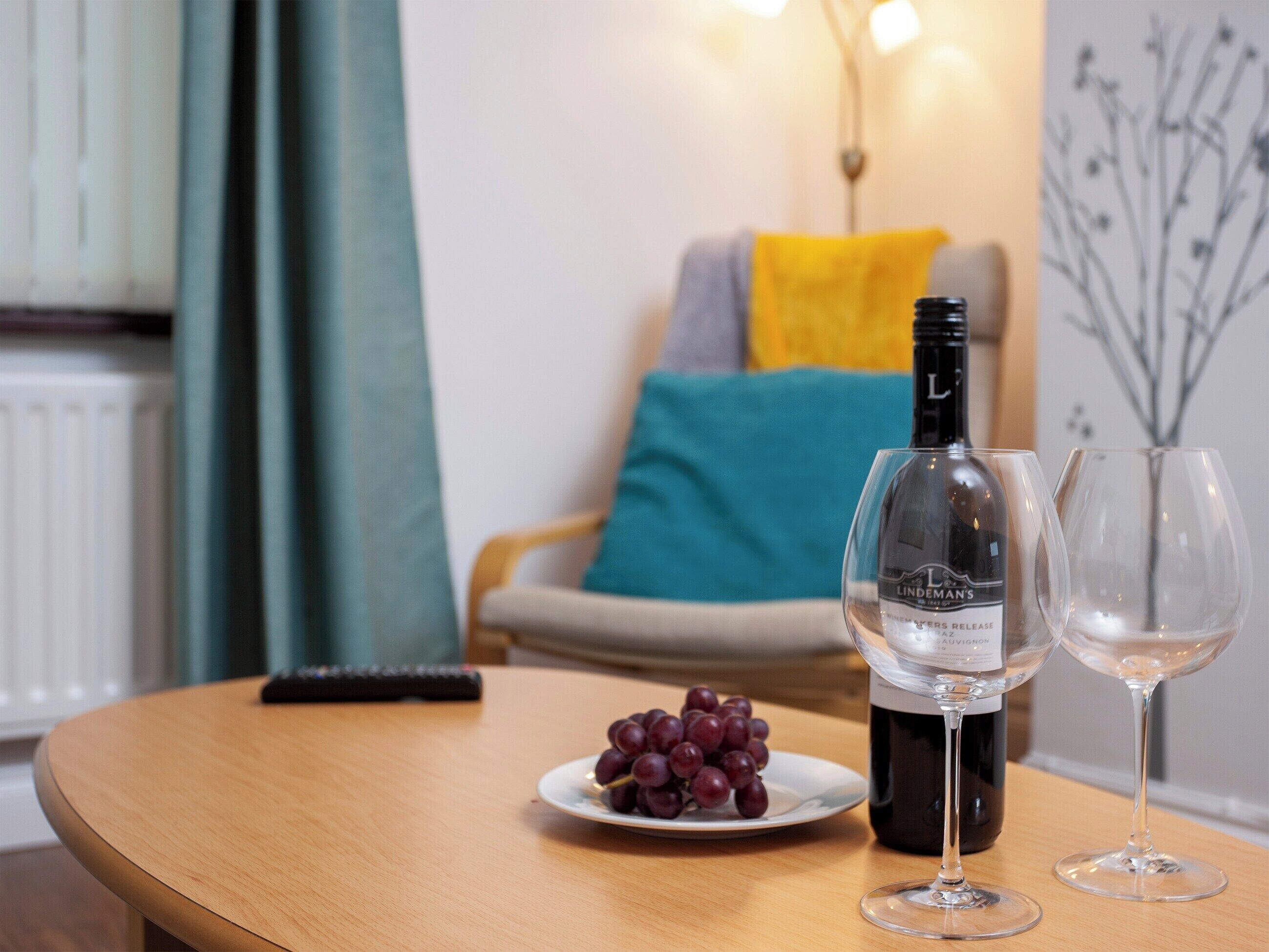 En flaska Lindemans-vin, två vinglas och en tallrik med vindruvor på ett träbord i ett vardagsrum.