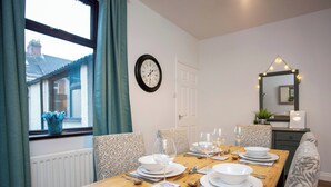 A dining room with a wooden table set for four, chairs with patterned cushions, a wall clock, a window with curtains, and a mirror with lights. - 2 bedroom accommodation in Shildon, near Bishop Auckland (Shildon, near Bishop Auckland)