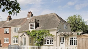 Une maison à deux étages, avec une clôture en bois peinte en blanc, une cheminée en briques et un jardin agrémenté de plantes suspendues.
