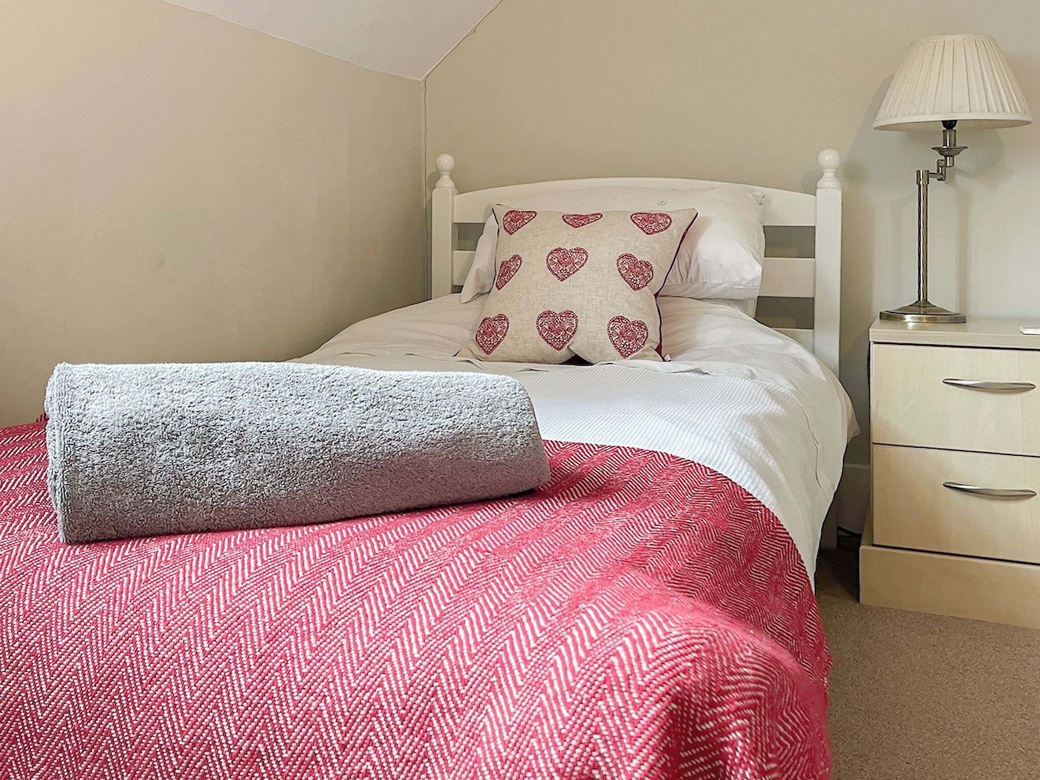 Una cama bien hecha con una colcha de cuadros rojos y blancos, una manta gris enrollada y almohadas con diseño de corazones. Se observa una mesita de noche con una lámpara y un mueble con cajones a la derecha.