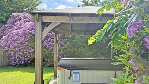 Een houten constructie met een bank en een hottub, omgeven door weelderige begroeiing en paarse bloemen.