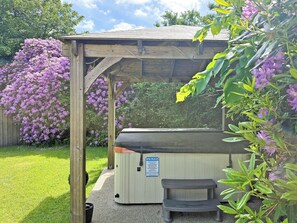 Une structure en bois abritant un banc et un jacuzzi, le tout entouré d’une végétation luxuriante et de fleurs violettes.