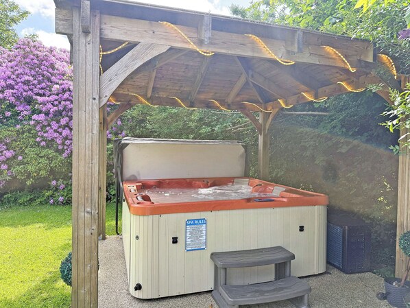 A hot tub with a wooden pergola overhead, surrounded by lush greenery and purple flowers.